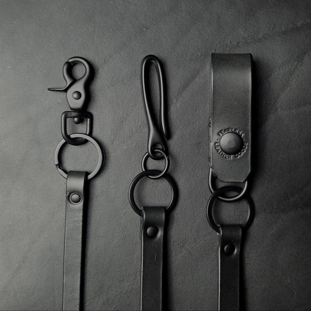 Three black leather strap, biker wallet chain with solid brass hardware. The left item has a swivel snap hook and ring, the middle has a fish hook and ring, and the right item has a leather belt loop with loop and ring.